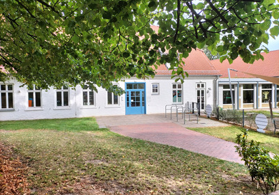 Ein helles Gebäude mit weißen Wänden und blauen Türen, umgeben von grünem Gras und Laubbäumen. Der Eingangsbereich ist gepflastert, und mehrere Fenster sind sichtbar. Im Hintergrund sieht man einen modernen Anbau. Das Gesamtbild vermittelt eine einladende Atmosphäre.