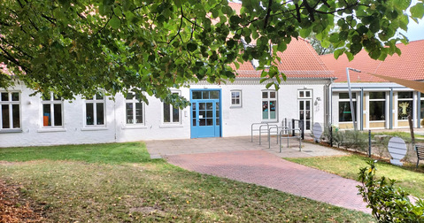 Ein helles Gebäude mit weißen Wänden und blauen Türen, umgeben von grünem Gras und Laubbäumen. Der Eingangsbereich ist gepflastert, und mehrere Fenster sind sichtbar. Im Hintergrund sieht man einen modernen Anbau. Das Gesamtbild vermittelt eine einladende Atmosphäre.
