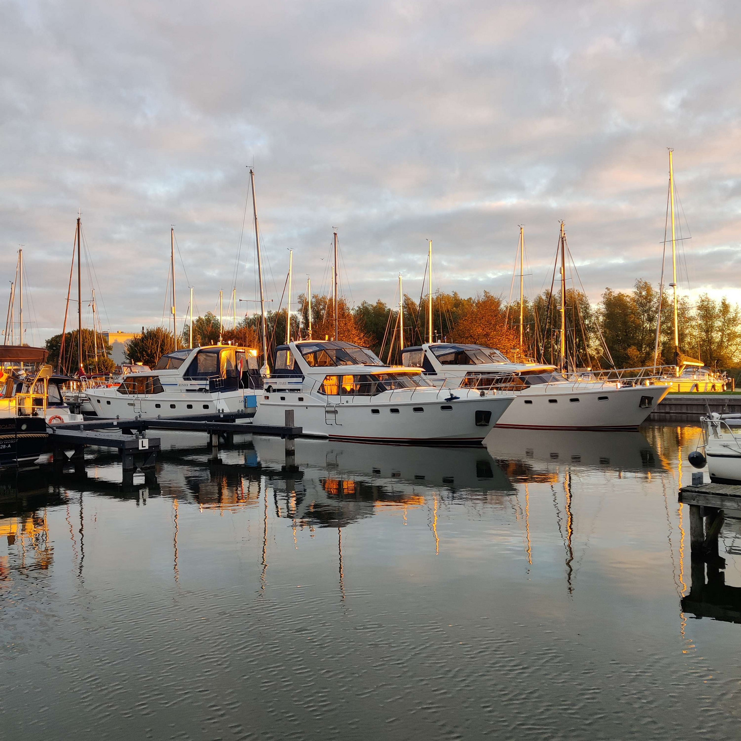 Sloten Morgens Sicht auf einen Hafen mit mehreren weißen Yachten, die im Wasser liegen. Die Mastspitzen ragen in den Himmel, und die sanfte Spiegelung der Boote zeigt sich im ruhigen Wasser. Der Himmel ist leicht bewölkt und es herrscht sanftes Licht.