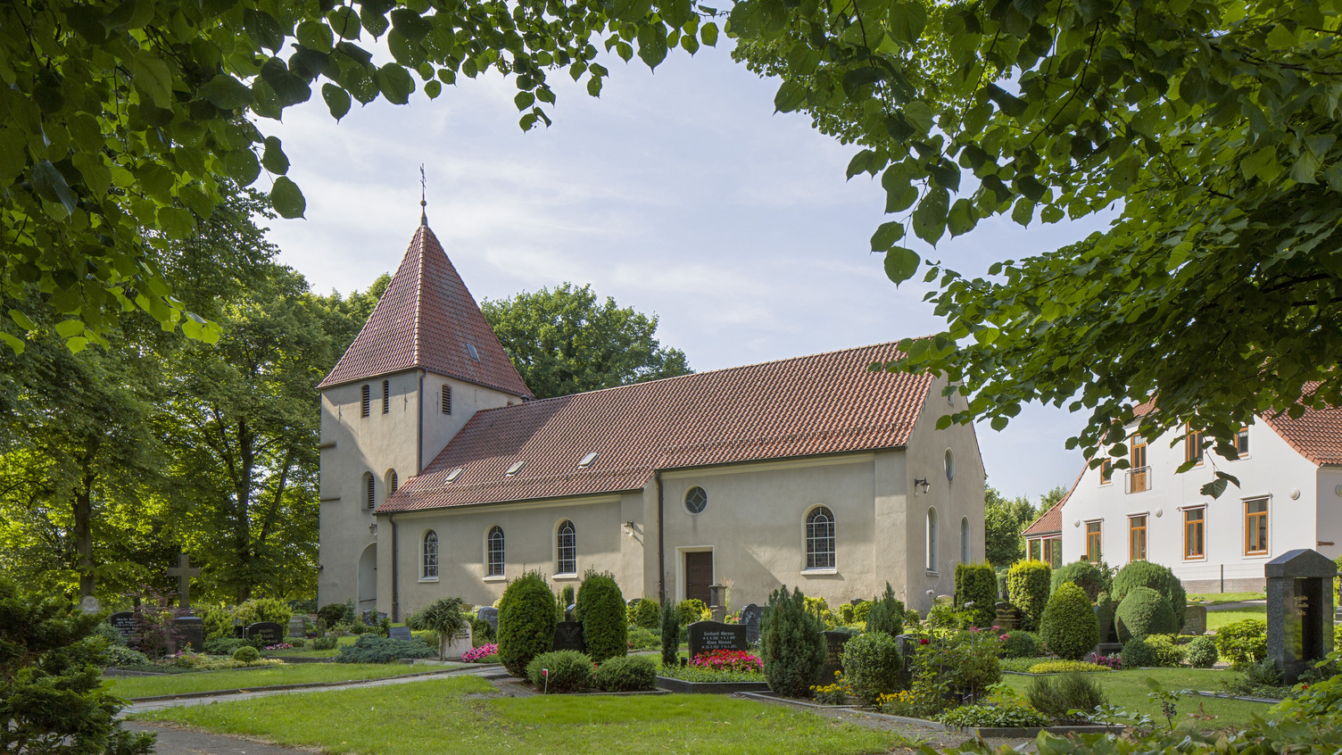 Eine einfache Kirche mit einem roten Satteldach und einem kleinen Turm steht umgeben von Bäumen und einem gepflegten Garten. Im Vordergrund sind Gräber mit Blumen und Sträuchern angeordnet, während im Hintergrund ein weiteres Gebäude zu erkennen ist.