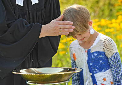 Das Foto zeigt die open air Taufe eines kleinen Jungen. Die Hand der Pastorin liegt auf seinem Kopf. Im Vordergrund steht das Messingtaufbecken, im Hintergrund sieht man gelb blühende Sträucher