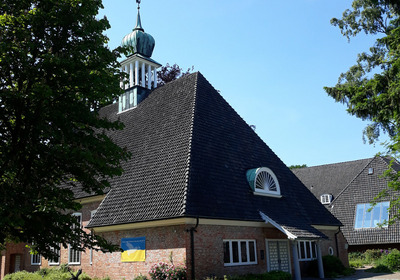 Ein Gebäude mit einem auffälligen, spitzen Dach und einer grünen Kirchturmspitze steht im Vordergrund. An der Wand des Gebäudes hängt eine Flagge der Ukraine. Um das Gebäude herum sind Bäume und Blumen zu sehen, unter blue skies.