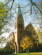Eine Backsteinkirche mit einem hohen, spitzen Turm ist im Bild zu sehen. Die Fassade zeigt eine Mischung aus Fenstern und architektonischen Details. Um die Kirche herum wachsen Bäume mit gelben Blättern, die auf einen herbstlichen Tag hindeuten.
