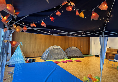Im Raum sind zwei Zeltstrukturen und ein farbiger Teppich aus Kissen. Oben hängen Lichterketten mit bunten Lampen. Ein blauer Mattenbereich ist deutlich sichtbar, neben dem befinden sich Spielzeug und bunte Bälle in einem Korb.