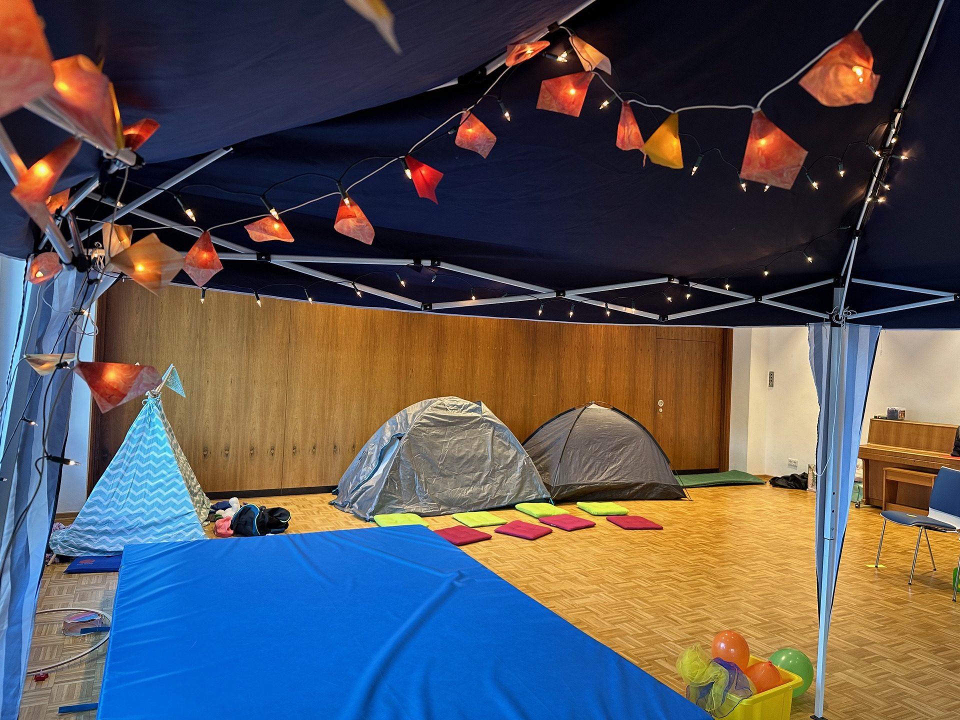 Im Raum sind zwei Zeltstrukturen und ein farbiger Teppich aus Kissen. Oben hängen Lichterketten mit bunten Lampen. Ein blauer Mattenbereich ist deutlich sichtbar, neben dem befinden sich Spielzeug und bunte Bälle in einem Korb.