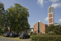 Ein moderner Kirchenbau aus rotem Ziegel mit einem hohen, schmalen Kirchturm, auf dem ein Kreuz steht. Vor der Kirche sind mehrere Autos geparkt, umgeben von Bäumen und grüner Vegetation. Der Himmel ist blau mit einigen Wolken.