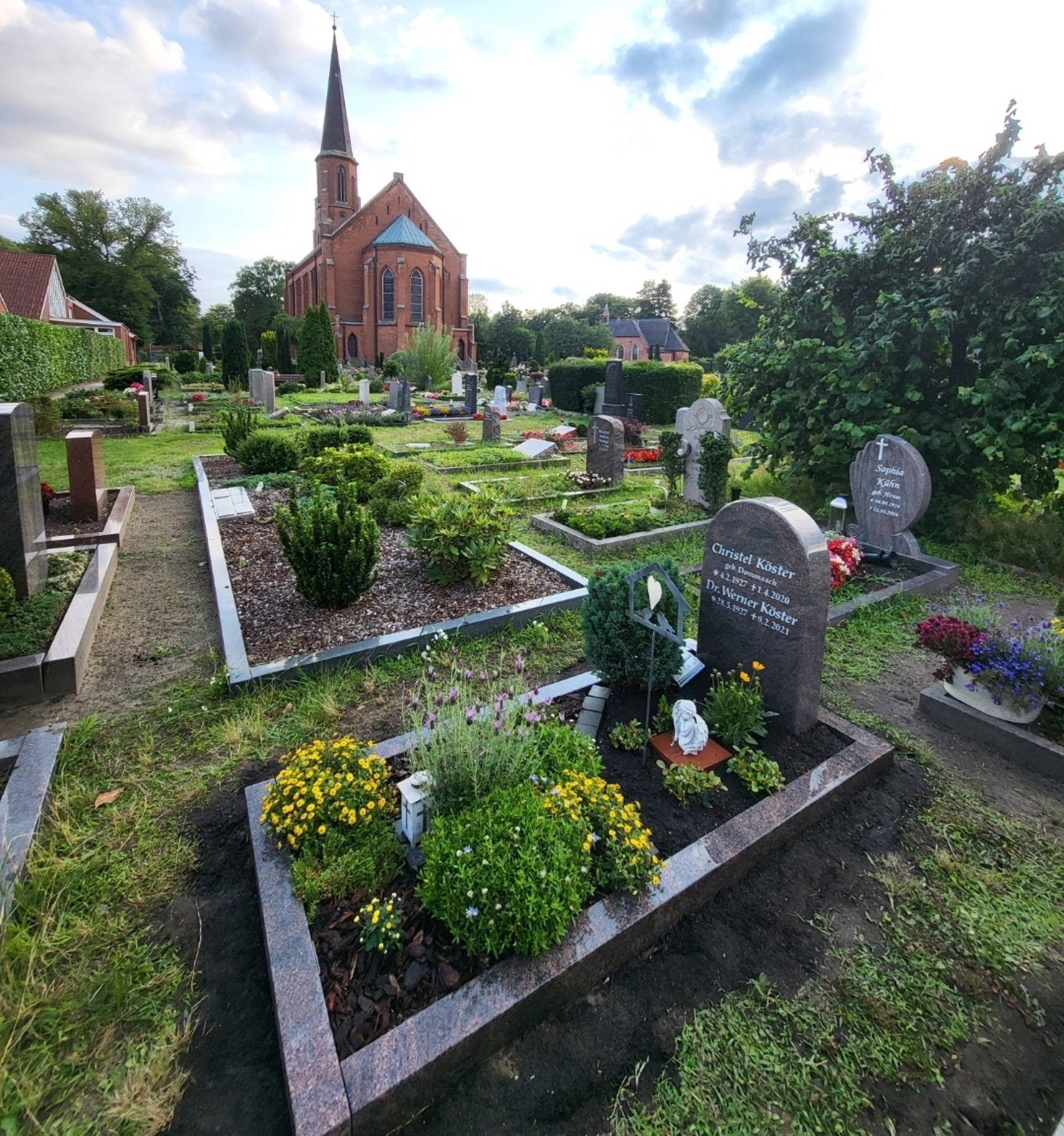 Ein Friedhof mit mehreren Grabstätten, geschmückt mit bunten Blumen und grünen Pflanzen. Im Hintergrund steht eine große, rote Kirche mit einem hohen Turm. Die Atmosphäre ist ruhig und respektvoll.
