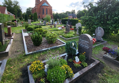 Ein Friedhof mit mehreren Grabstätten, geschmückt mit bunten Blumen und grünen Pflanzen. Im Hintergrund steht eine große, rote Kirche mit einem hohen Turm. Die Atmosphäre ist ruhig und respektvoll.