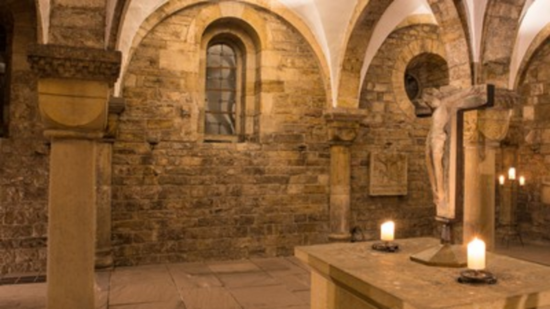Eine historische Kapelle mit steinernen Wänden und gewölbten Decken. In der Mitte steht ein Tisch mit Kerzen und einer Kreuzigungsszene. Die gedämpfte Beleuchtung schafft eine ruhige, spirituelle Atmosphäre.