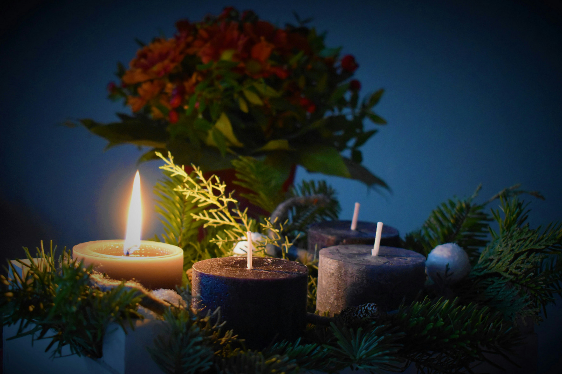 Ein Arrangement aus vier Kerzen, von denen eine brennt, umgeben von Tannenzweigen und Dekor. Im Hintergrund sind farbige Blumen sichtbar. Die Szene vermittelt eine gemütliche, festliche Atmosphäre.