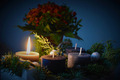 Ein Arrangement aus vier Kerzen, von denen eine brennt, umgeben von Tannenzweigen und Dekor. Im Hintergrund sind farbige Blumen sichtbar. Die Szene vermittelt eine gemütliche, festliche Atmosphäre.