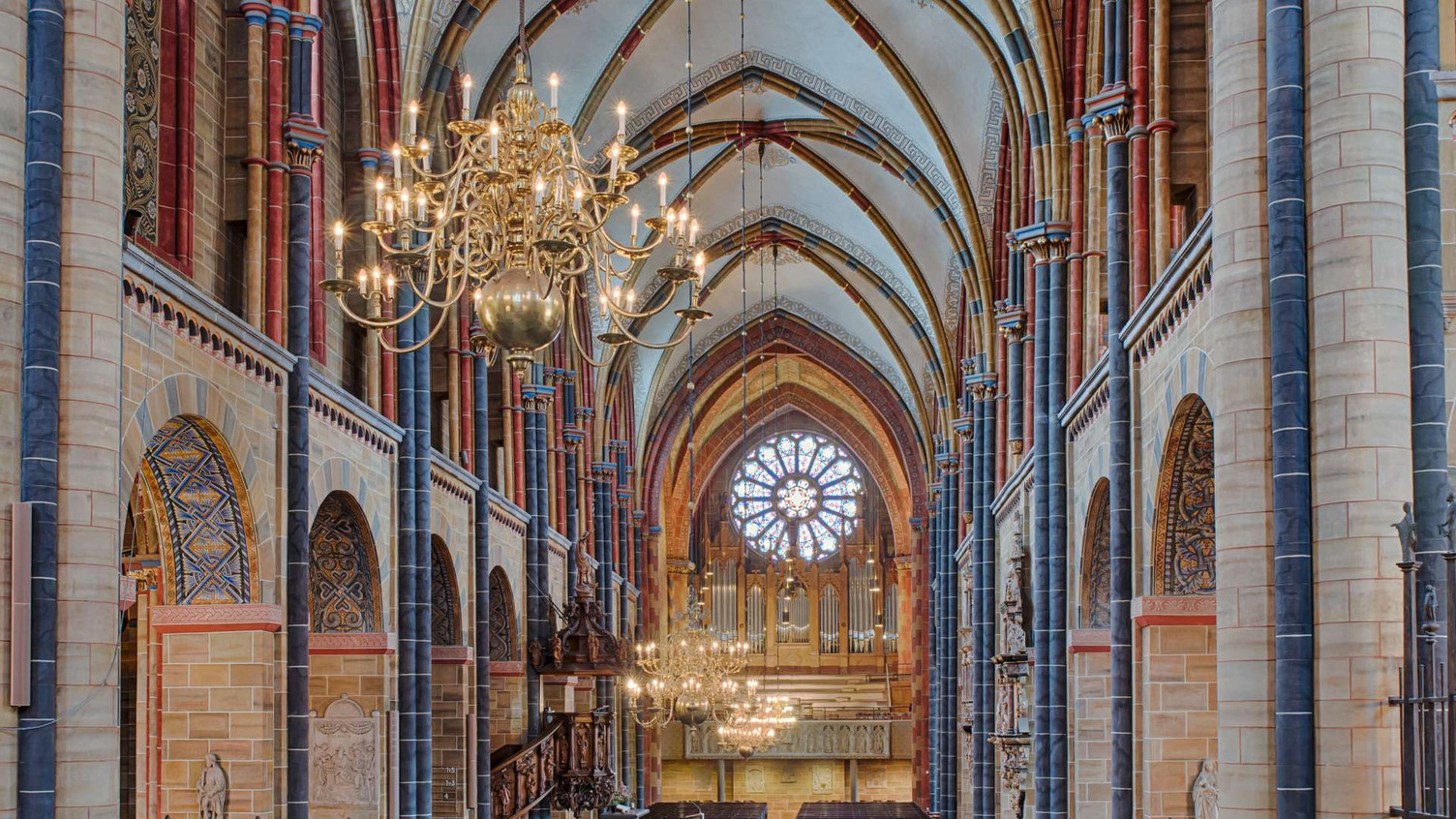 Blick ins Innere einer beeindruckenden Kirche mit hohen, gewölbten Decken und einer zentralen, kunstvollen Fensterrose. Mehrere massive Säulen flankieren den Raum, der von einer großen, dekorativen Deckenleuchte erhellt wird. Im Hintergrund sind Altäre und religiöse Kunstwerke zu erkennen.