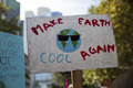 Ein Schild bei einer Demonstration zeigt die Erde mit Sonnenbrille und den Worten "MAKE EARTH COOL AGAIN". Im Hintergrund sind verschwommene Gebäude und Bäume zu sehen.