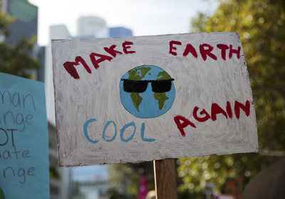 Ein Schild bei einer Demonstration zeigt die Erde mit Sonnenbrille und den Worten "MAKE EARTH COOL AGAIN". Im Hintergrund sind verschwommene Gebäude und Bäume zu sehen.