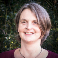 Eine lächelnde Frau mit schulterlangen braunen Haaren trägt ein dunkelrotes Oberteil und eine Halskette. Sie steht vor einer grünen, unscharfen Hintergrundlandschaft, die etwas Natur zeigt.