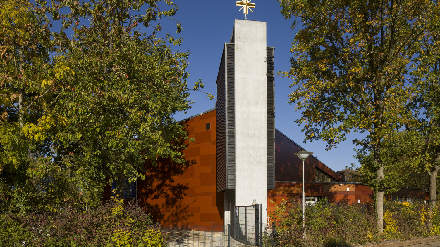 Ein modernes Kirchengebäude mit einer orangefarbenen Fassade und einem schlichten, hohen Turm. Auf dem Turm befindet sich ein goldenes Kreuz. Rund um das Gebäude sind Bäume und Sträucher mit bunten Blättern, die eine herbstliche Atmosphäre vermitteln. Der Himmel ist klar und blau.