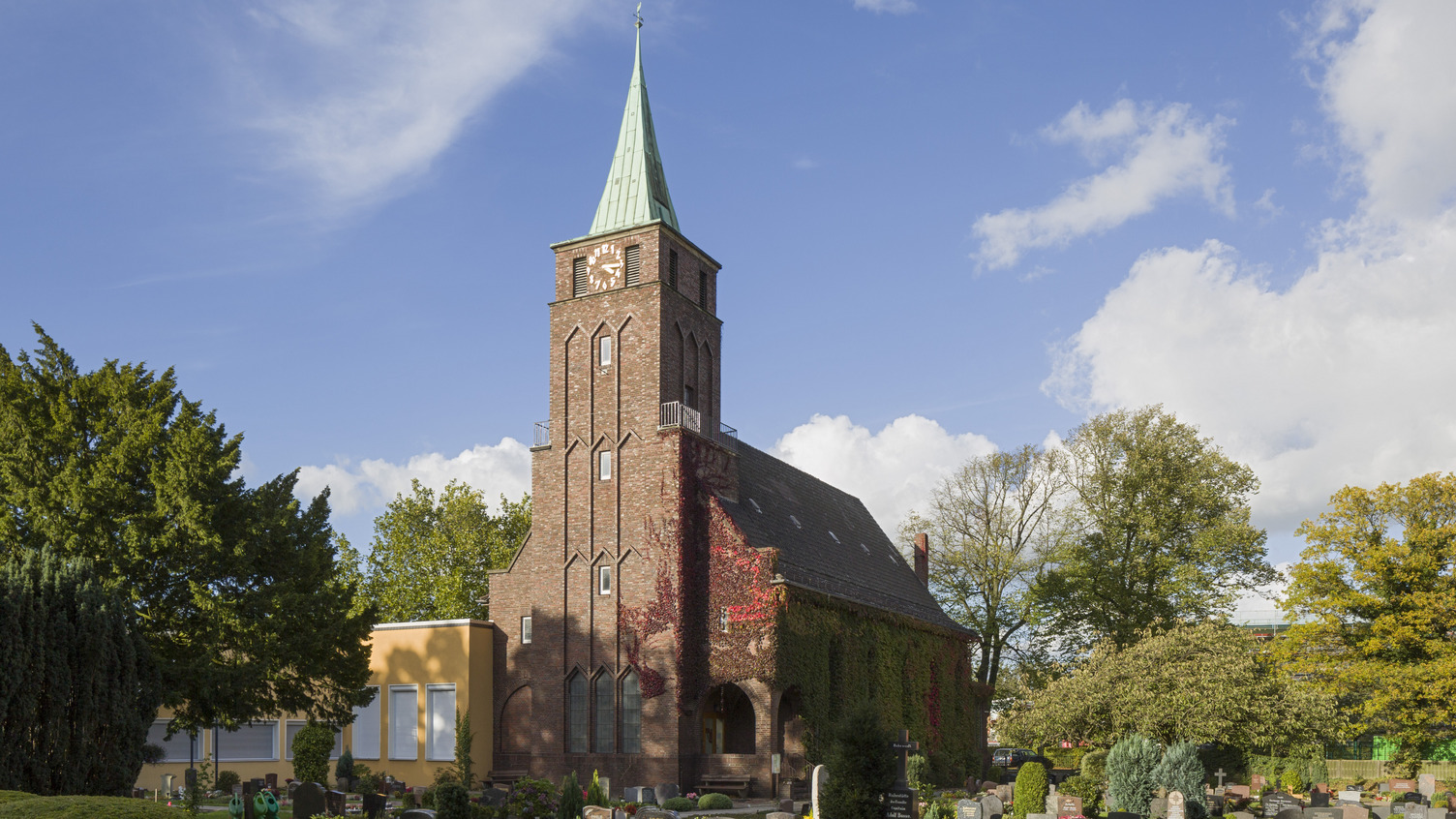 Eine kleine, gemauerte Kirche mit einem hohen, spitzen Turm, der eine grüne Kupferdachspitze hat. Umgeben von einem Friedhof mit Grabsteinen und grünen Bäumen, unter einem klaren, blauen Himmel mit einigen Wolken.