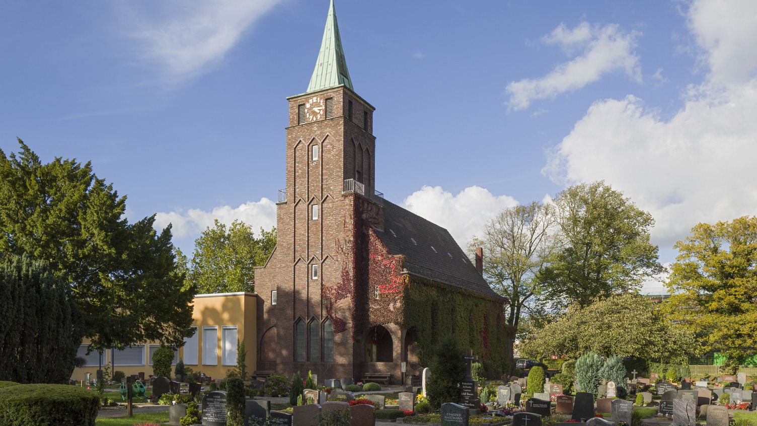 Eine kleine, gemauerte Kirche mit einem hohen, spitzen Turm, der eine grüne Kupferdachspitze hat. Umgeben von einem Friedhof mit Grabsteinen und grünen Bäumen, unter einem klaren, blauen Himmel mit einigen Wolken.