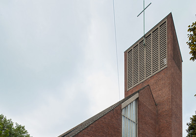 Eine moderne, aus roten Ziegeln errichtete Kirche mit einem markanten Turm. Der Turm hat ein Kreuz an der Spitze und große Fenster im unteren Bereich. Der Himmel ist bewölkt, und im Vordergrund sind teilweise Bäume sichtbar.