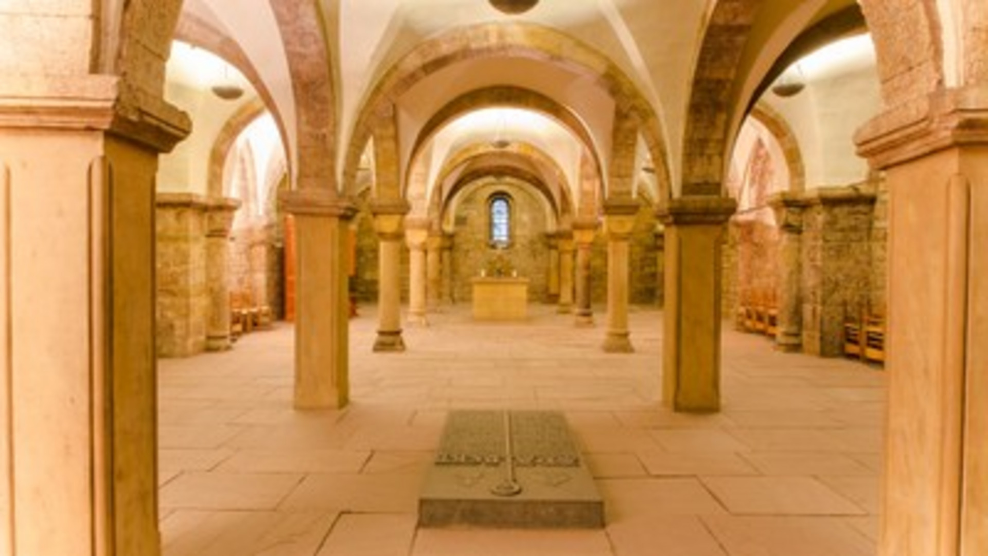 Der Innenraum einer historischen Krypta mit hohen Bögen und Säulen. In der Mitte befindet sich ein schlichter Altar, und am Boden liegt ein Gedenkstein. Sanftes Licht fällt durch die Fenster, und die Wände sind aus hellem Stein. Die Atmosphäre ist ruhig und ehrwürdig.