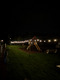 Ein Spielplatz bei Nacht, beleuchtet von warmen Lichtern. Im Vordergrund sieht man eine Rutsche und Kletterstruktur aus Holz, umgeben von grünem Rasen. Der Hintergrund ist dunkel, mit der Silhouette von Bäumen. Links ist ein Steg zu sehen, rechts steht ein Wohnmobil.