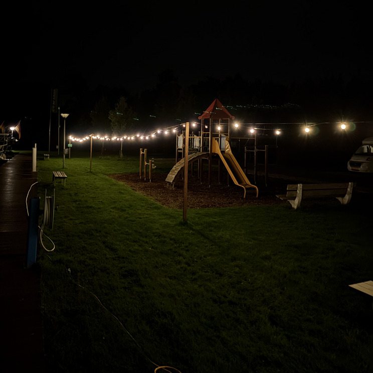Sneek Lichterkette Ein Spielplatz bei Nacht, beleuchtet von warmen Lichtern. Im Vordergrund sieht man eine Rutsche und Kletterstruktur aus Holz, umgeben von grünem Rasen. Der Hintergrund ist dunkel, mit der Silhouette von Bäumen. Links ist ein Steg zu sehen, rechts steht ein Wohnmobil.