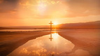 Ein kreuzförmiger Pfahl steht inmitten einer ruhigen Landschaft, während die Sonne aufgeht und den Himmel in warmen Farben erleuchtet. Ein Wasserbecken spiegelt das Licht und die Umgebung wider, was eine friedliche und spirituelle Atmosphäre schafft.