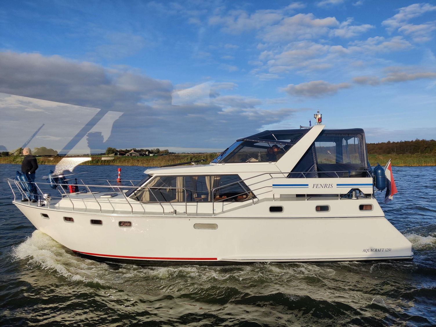 Fenris PM Eine weiße Yacht mit dem Namen "Fenris" fährt auf einem Gewässer. Der Pastor steht an der Vorderseite des Bootes und aus dem Steuerstand ragen der Kopf der Kapitänin und einer Teamerin hervor. Im Hintergrund sind grüne Uferlandschaften und ein blauer Himmel mit Wolken sichtbar.