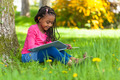Auf dem Foto ist ein kleines Mädchen zu sehen, das auf einer Wiese unter einem Baum sitzt und in einem Bilderbuch liest