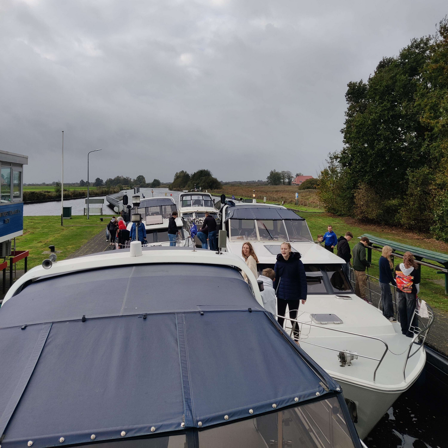 Homansluis Vier Boote liegen in einer Schleuse. Personen stehen auf den Decks und am Ufer, einige unterhalten sich oder bewegen sich umher. Der Himmel ist bewölkt, und die umgebende Landschaft besteht aus Bäumen und Wiesen.