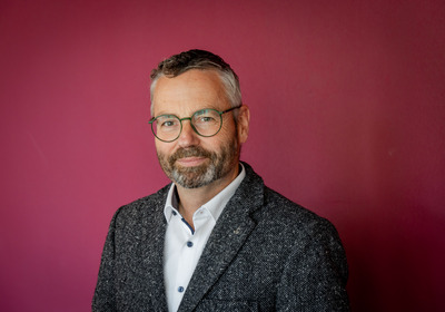 Ein Mann mit dunklen Haaren und einem Bart trägt eine Brille. Er hat ein freundliches Lächeln und ist in einer grauen Jacke und einem weißen Hemd gekleidet. Der Hintergrund ist in einem dunklen Pinkton gehalten.