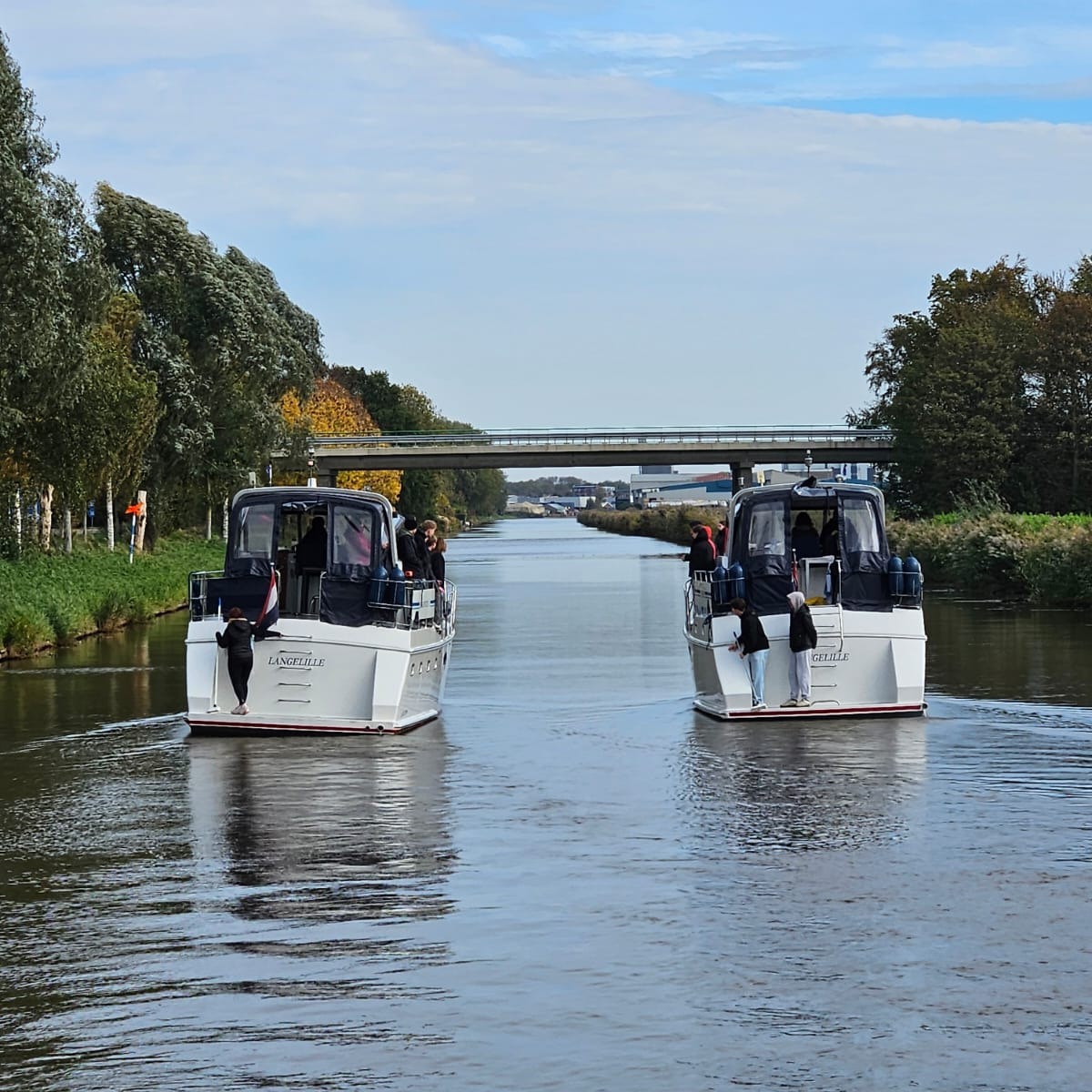Ballspielen Zwei Boote fahren auf einem ruhigen Wasserweg. Personen stehen auf den Decks und schauen in verschiedene Richtungen. Sie werfen sich einen Ball zu, vom einen Boot auf das andere. Im Hintergrund ist eine Brücke sichtbar und die Ufer sind von Bäumen gesäumt. Der Himmel ist leicht bewölkt.