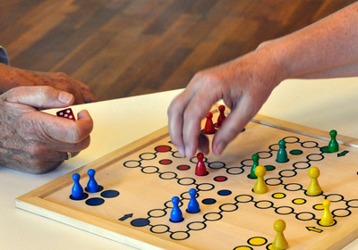 Zwei Hände über einem Mensch-ärgere-dich-nicht-Brett, eine zieht eine Spielfigur, die andere hält einen Würfel