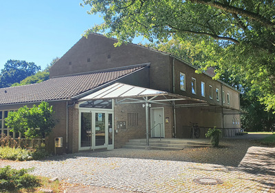 Ein graues Backsteingebäude mit schrägen Dächern und einem überdachten Eingangsbereich an einem sonnigen Tag.