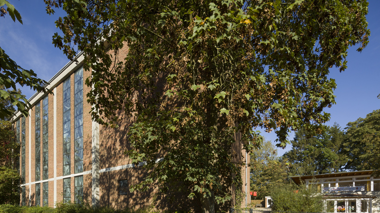 Ein Gebäude mit roten Ziegeln und großen Fenstern ist von Bäumen und Sträuchern umgeben. Der Himmel ist klar und blau, und ein Weg führt zu einem weiteren, hellen Gebäude auf der rechten Seite.
