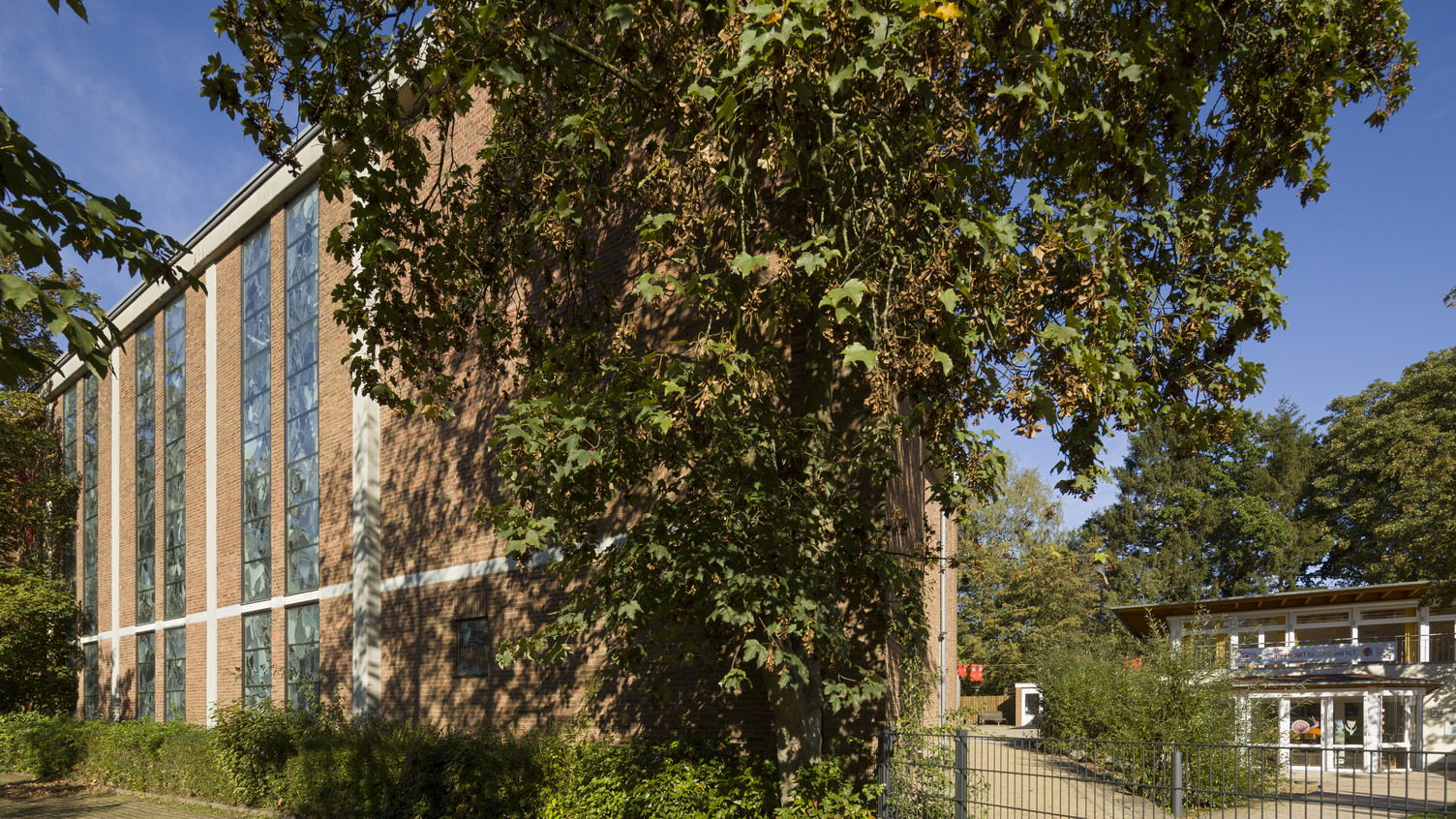 Ein Gebäude mit roten Ziegeln und großen Fenstern ist von Bäumen und Sträuchern umgeben. Der Himmel ist klar und blau, und ein Weg führt zu einem weiteren, hellen Gebäude auf der rechten Seite.