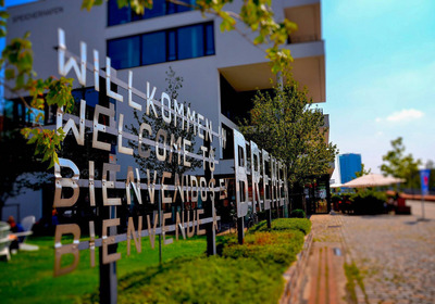 Ein transparentes Schild mit der Aufschrift „Willkommen in Bremen“ in mehreren Sprachen steht vor einem modernen Gebäude. Umgeben von Bäumen und Grünflächen bildet die Szene einen einladenden Eingangsbereich.