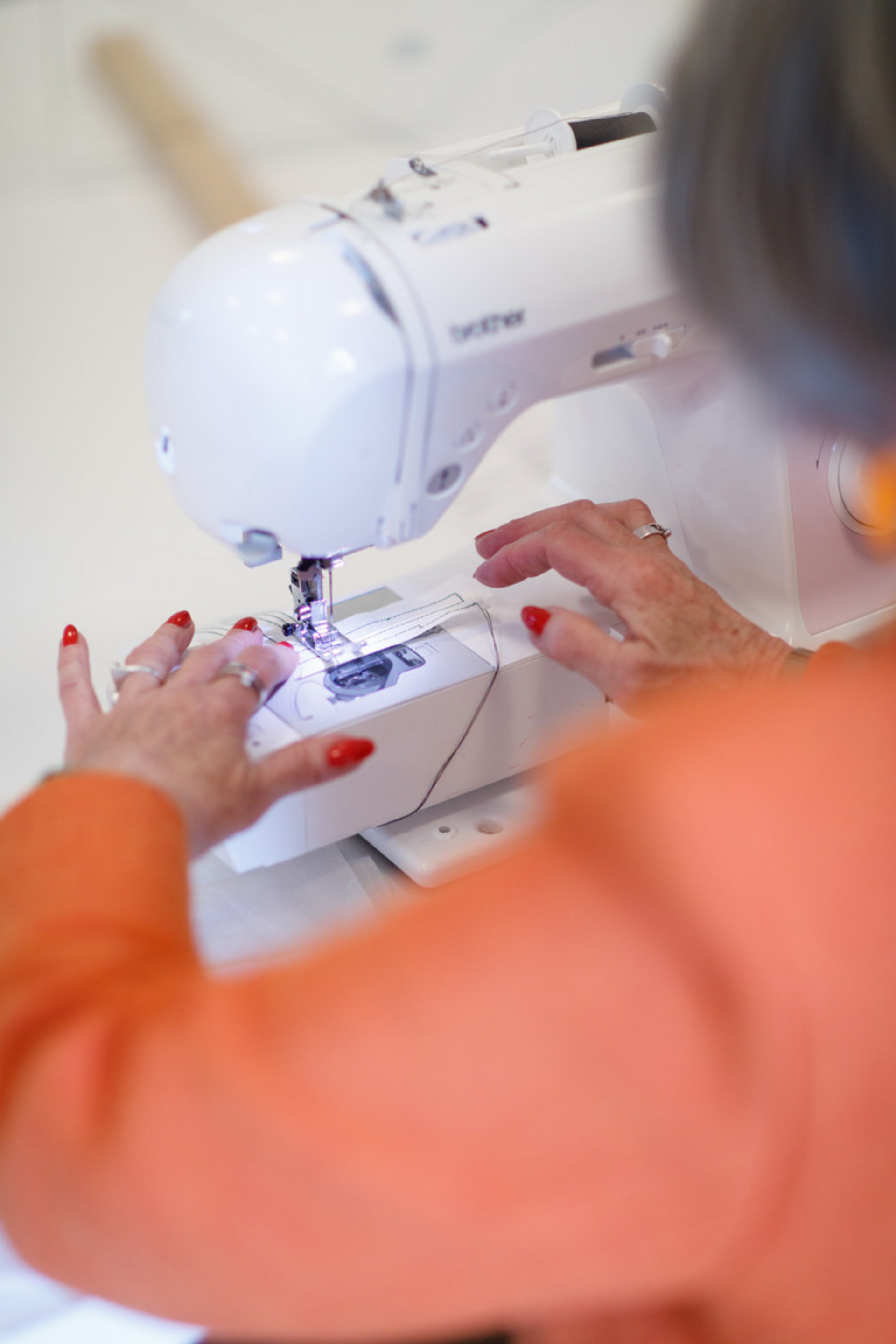 Eine Person mit einem orangenen Oberteil näht an einer Nähmaschine. Ihre Hände sind sichtbar, während sie den Stoff führt. Die Maschine ist modern und auf einem Tisch platziert, umgeben von einem hellen Arbeitsbereich.