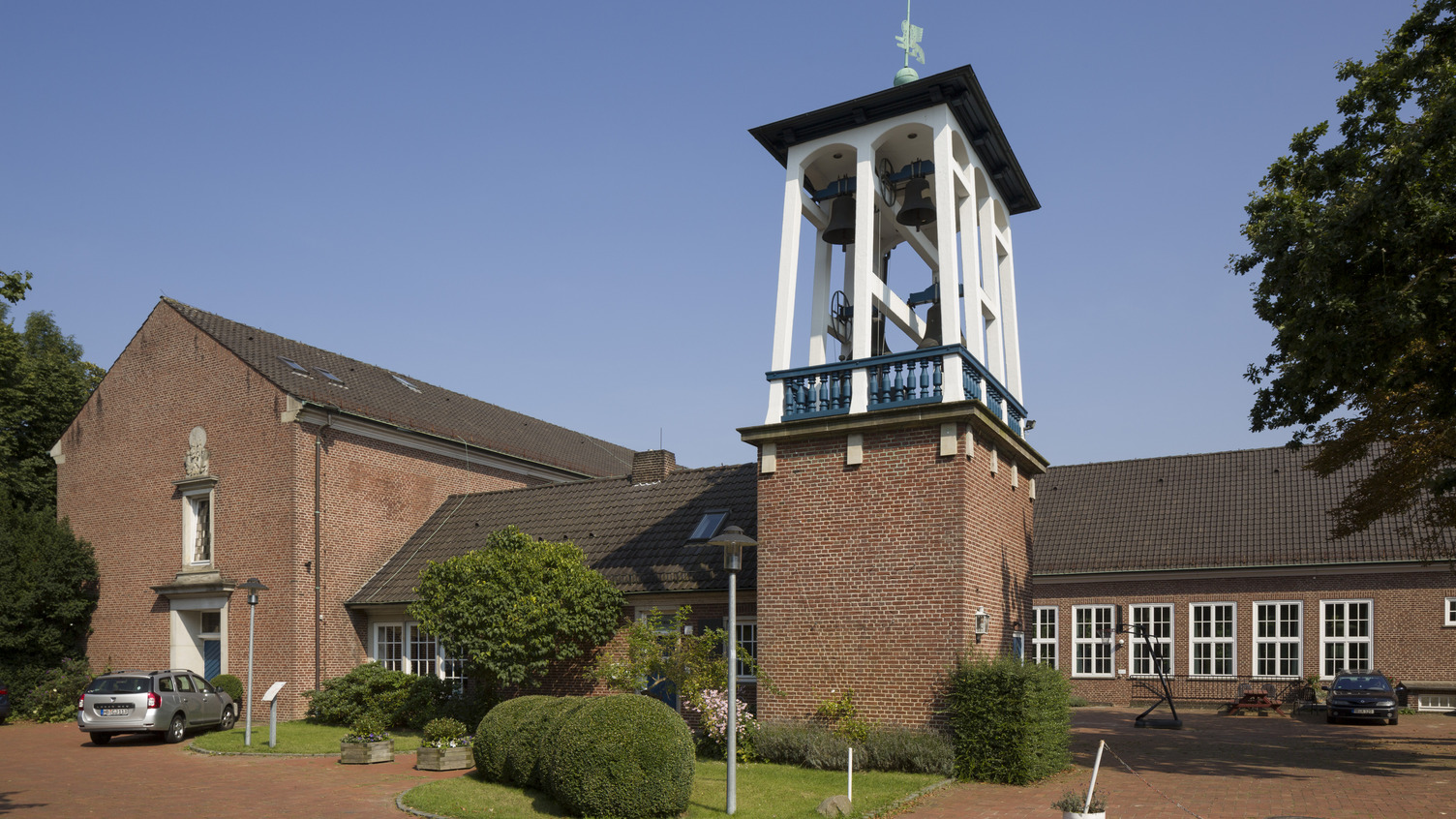 Ein rotes Backsteingebäude mit einem schlichten, weißen Glockenturm und mehreren Fenstern. Umgeben von grünen Pflanzen und einem gepflasterten Vorplatz. Der Himmel ist blau und wolkenlos.