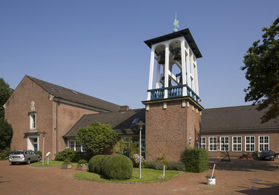 Ein rotes Backsteingebäude mit einem schlichten, weißen Glockenturm und mehreren Fenstern. Umgeben von grünen Pflanzen und einem gepflasterten Vorplatz. Der Himmel ist blau und wolkenlos.