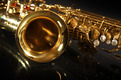 Ein goldfarbenes Saxophon liegt auf einem dunklen Untergrund, wobei der Blick auf den Schallbecher und einige Klappen fällt. Die glänzende Oberfläche reflektiert das Licht und hebt die Details des Instruments hervor.