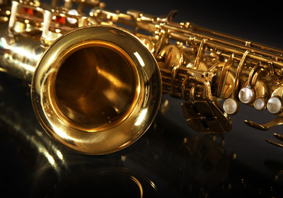 Ein goldfarbenes Saxophon liegt auf einem dunklen Untergrund, wobei der Blick auf den Schallbecher und einige Klappen fällt. Die glänzende Oberfläche reflektiert das Licht und hebt die Details des Instruments hervor.
