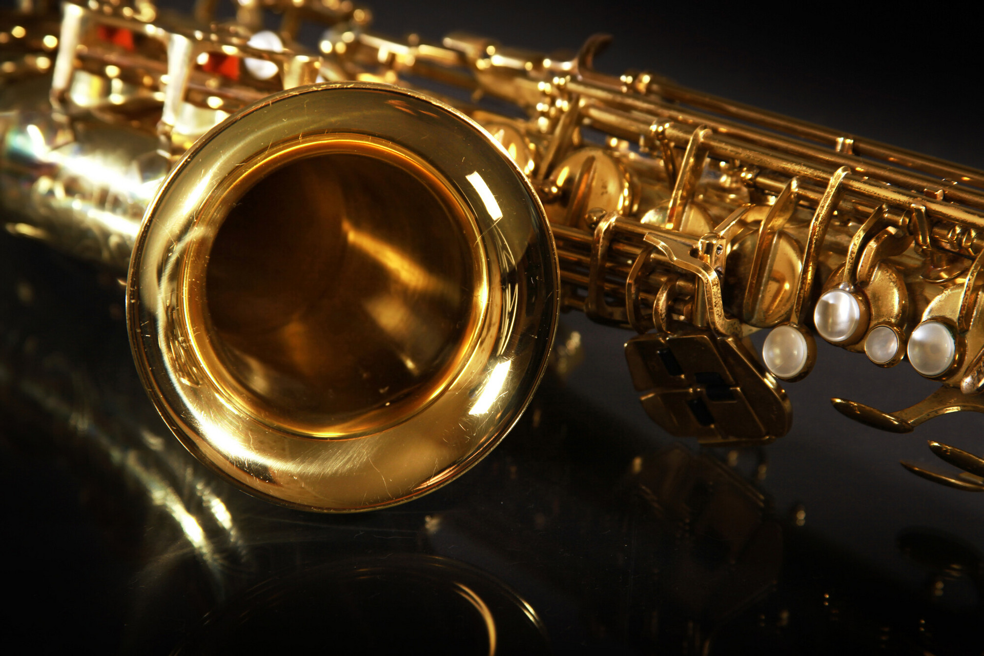 Ein goldfarbenes Saxophon liegt auf einem dunklen Untergrund, wobei der Blick auf den Schallbecher und einige Klappen fällt. Die glänzende Oberfläche reflektiert das Licht und hebt die Details des Instruments hervor.
