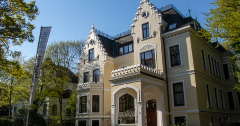 Eine elegante, historische Villa mit hellgelber Fassade und kunstvollen Details. Die Gebäude besitzen mehrere Fenster und ein dekoratives Dach. Vor dem Haus sind grüne Bäume und ein gepflegter Garten sichtbar, unter einem klaren blauen Himmel.