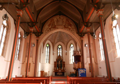 Der Innenraum einer Kirche mit einem hohen, gewölbten Decke. Holzbänke stehen in Reihen. Im Hintergrund befindet sich ein Altar mit einem aufwendigen Kanzel. Bunte Fenster und dekorative Malereien schmücken die Wände und verleihen dem Raum eine ruhige, spirituelle Atmosphäre.