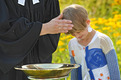 Das Foto zeigt die open air Taufe eines kleinen Jungen. Die Hand der Pastorin liegt auf seinem Kopf. Im Vordergrund steht das Messingtaufbecken, im Hintergrund sieht man gelb blühende Sträucher