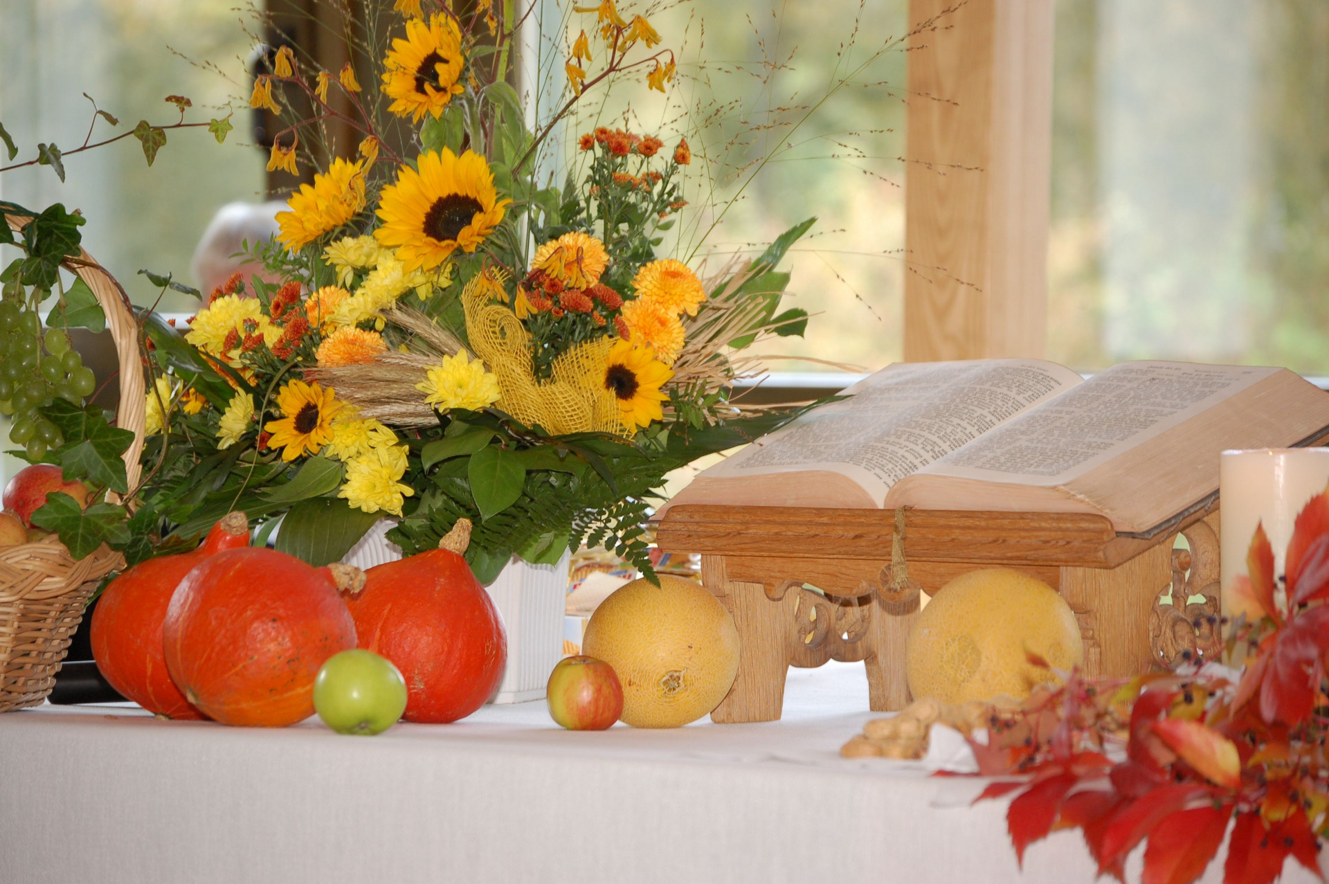 Ein Tisch mit einem offenen Buch auf einem Holzständer. Daneben liegen farbenfrohe Kürbisse, verschiedene Früchte wie Äpfel und Trauben sowie ein üppiger Blumenstrauß mit Sonnenblumen und weiteren Blüten. Die Szene strahlt eine herbstliche und festliche Atmosphäre aus.