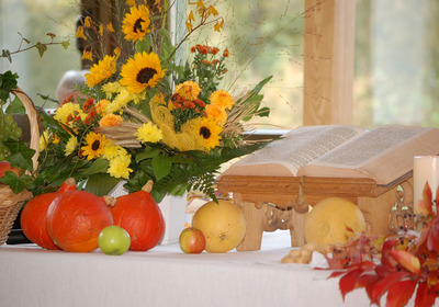 Ein Tisch mit einem offenen Buch auf einem Holzständer. Daneben liegen farbenfrohe Kürbisse, verschiedene Früchte wie Äpfel und Trauben sowie ein üppiger Blumenstrauß mit Sonnenblumen und weiteren Blüten. Die Szene strahlt eine herbstliche und festliche Atmosphäre aus.