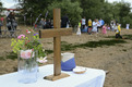 Ein kleiner Altar am Strand mit einem Holzkreuz un d Blumen. Im Hintergrund Taufgäste.