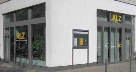 An der Ecke eines Gebäudes befindet sich ein Eingang mit großen Fenstern. Über dem Eingang steht das Logo „ALZ“ in gelben Buchstaben. Neben dem Eingang gibt es ein Info-Täfelchen mit verschiedenen Aushängen. Der Gehweg ist gepflastert und Palmgewächse sind sichtbar.